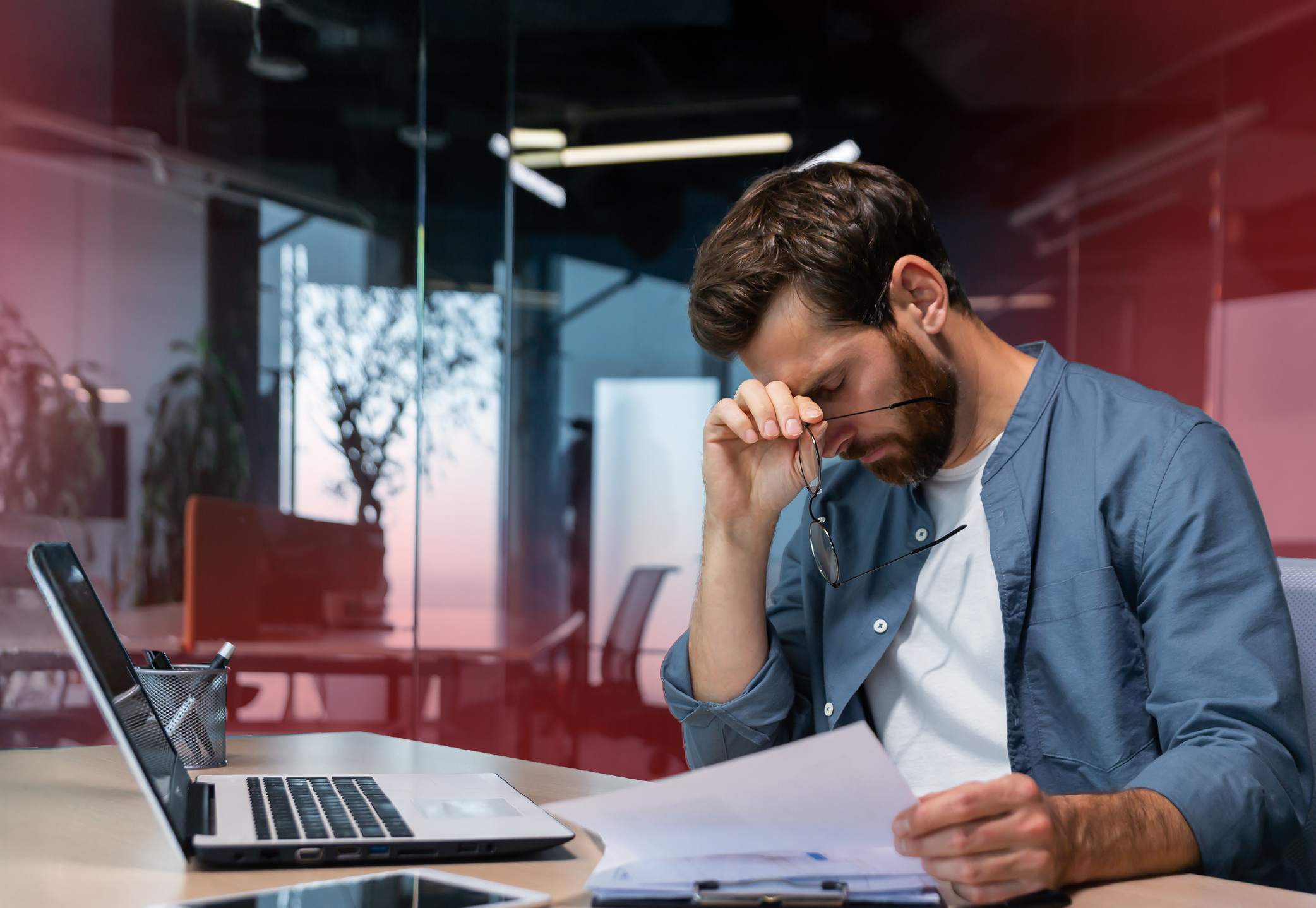 A man seated at a desk in an office environment, appearing stressed or fatigued as he pinches the bridge of his nose while holding a pair of glasses. A laptop, document, and pen holder are on the desk, with a modern office background featuring glass walls and plants. The subdued lighting and reddish tint add to the contemplative or emotional tone of the scene.