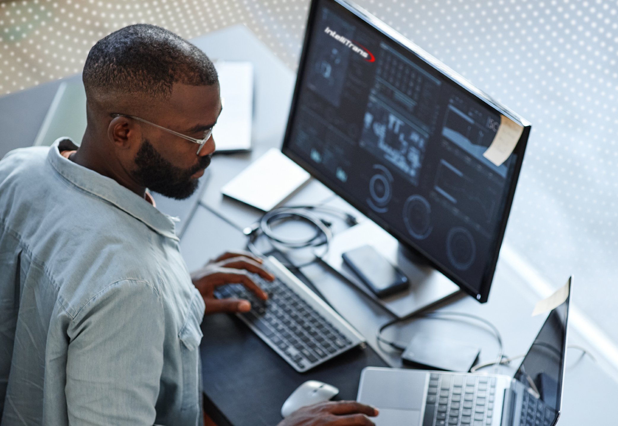 A person working at a desk with a large monitor displaying graphs, charts, and technical data. The setup includes a keyboard, mouse, and a partially visible laptop on the right. Sticky notes are attached to the monitor and laptop, and a smartphone and cables are on the desk. The scene suggests data analysis or technical monitoring in a professional office environment.
