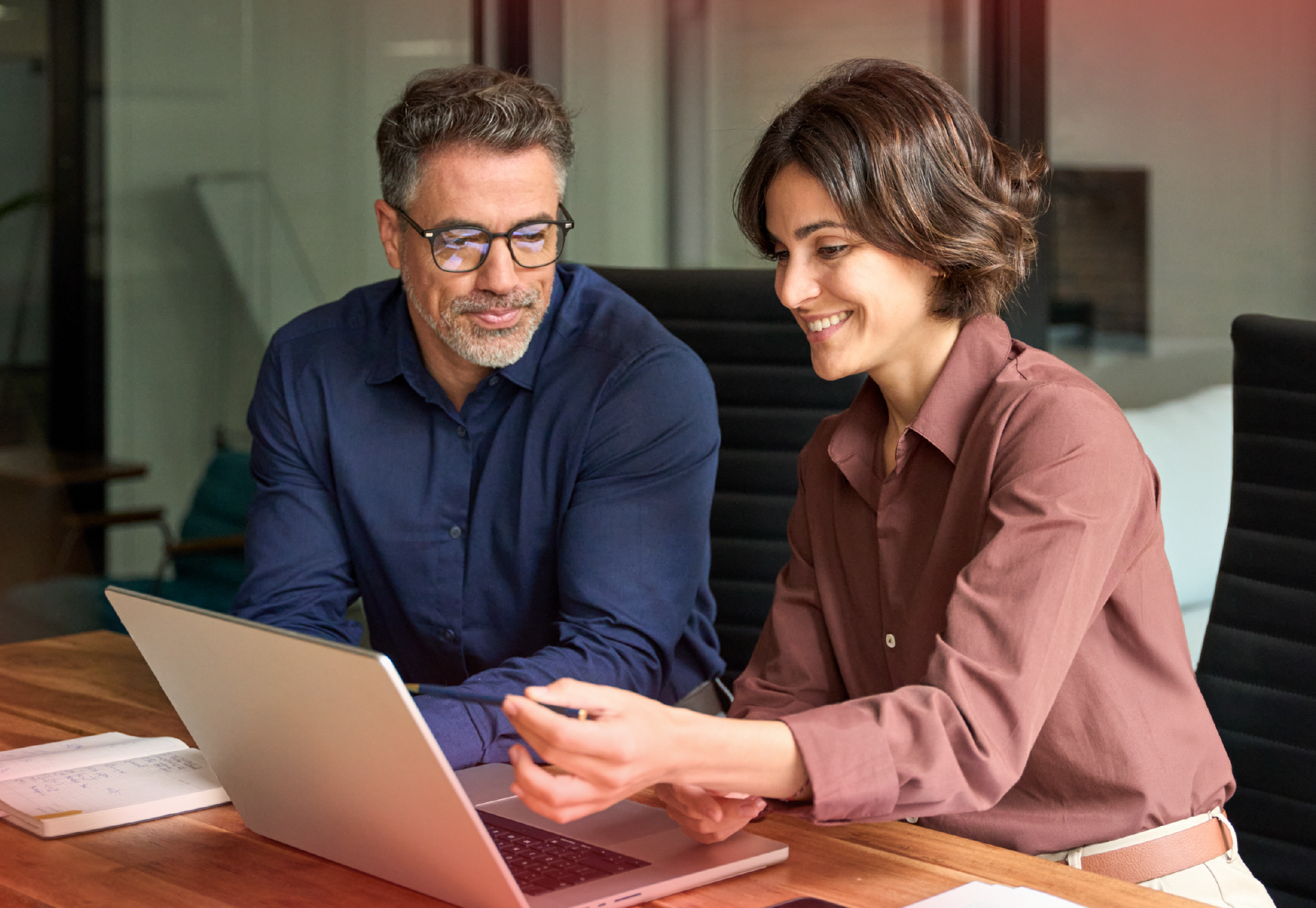 Two individuals seated at a table in a modern office setting, engaged in a collaborative discussion. One person gestures towards a laptop screen, while the other listens attentively. A notebook is placed on the table near the laptop, suggesting note-taking or data review. The background features a glass wall, adding to the professional and open environment.